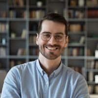 Hombre sonriente con gafas y camisa celeste, de pie frente a una estantería con libros, representando a un cliente satisfecho - testimonio cliente, confianza, profesionalismo, servicio web, experiencia positiva. | Diseño Web – SZ Web Design.