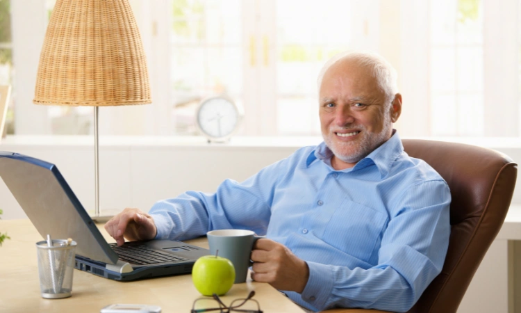 Hombre mayor sonriendo mientras trabaja con su laptop en una oficina iluminada.