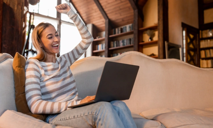 Mujer feliz usando su laptop en casa celebrando un logro en línea.