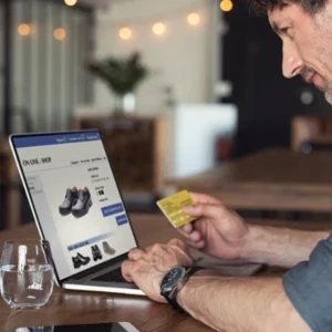 Hombre realizando compra online con tarjeta de crédito en su laptop en un ambiente moderno y acogedor.